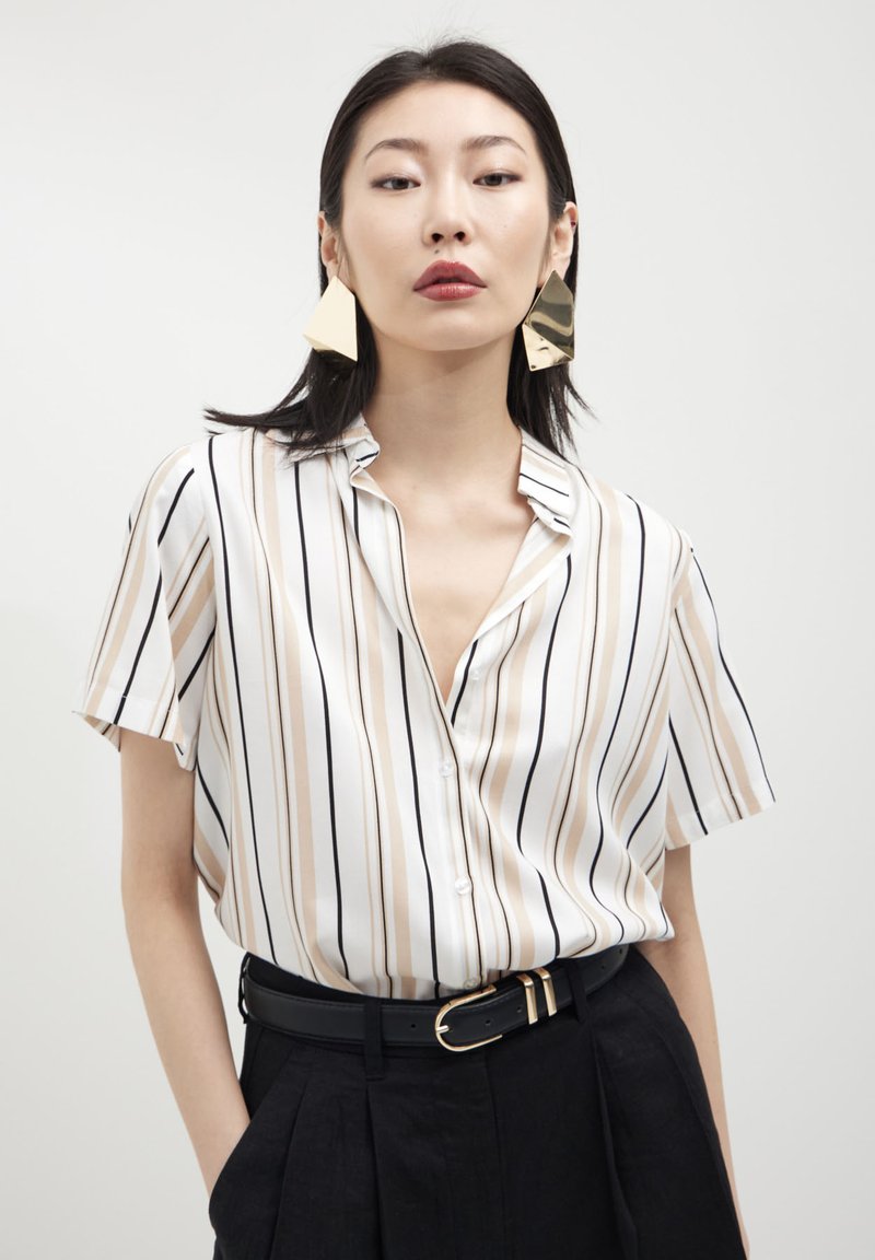 Camicia a maniche corte con colletto, a righe verticali beige e nere su uno sfondo bianco, abbinata a pantaloni neri a vita alta e una cintura nera.