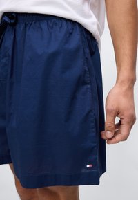 Άνδρας που φοράει σκούρο μπλε σορτς με ελαστική μέση και μικρό κόκκινο, άσπρο και μπλε λογότυπο κοντά στο τελείωμα, στέκεται μπροστά σε απλό φόντο.