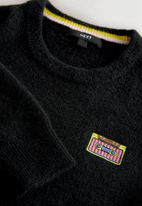 Pull en tricot noir avec une texture fuzzy, col rond et un motif de cassette audio coloré brodé sur le devant. Accents rayés vifs au niveau du col.