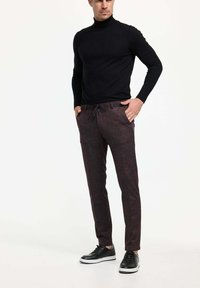 Dunkler Rollkragenpullover, enganliegendes Design. Dunkle gemusterte Hose mit Kordelzugbund. Schwarze Sneaker mit weißen Sohlen.