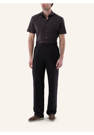 Mann trägt ein dunkelbraunes, kurzärmeliges Hemd mit Knöpfen, schwarze hochtaillierte Hose und braune Slipper auf hellem Hintergrund.