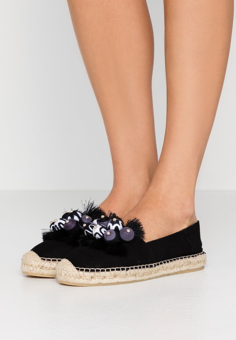 Espadrilles noires avec semelles en jute tissé, décorées de pompons noirs et blancs ainsi que de petites perles, portées pieds nus.