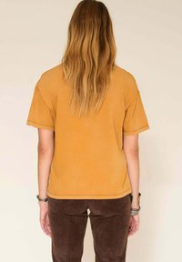 T-shirt décontracté de couleur moutarde avec manches courtes, coupe décontractée et détails de coutures visibles, associé à un pantalon slim marron.