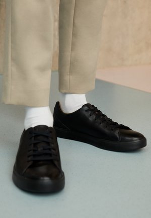Osoba nosząca beżowe spodnie, białe skarpetki i czarne skórzane trampki na sznurowadła stoi na jasnoniebieskiej podłodze.