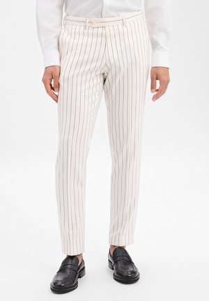Uomo che indossa pantaloni slim-fit color crema con sottili strisce verticali nere, abbinati a mocassini neri in pelle e una camicia bianca elegante.