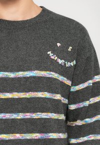 Mørkegrå strikkegenser med flerfargede horisontale striper og et lite fargerikt smilefjes-design på den øvre brystområdet.