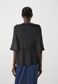 Blusa nera trasparente con collo e maniche arricciate, caratterizzata da un design peplum fluido, indossata sopra una gonna di jeans, vista da dietro.