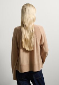 Beige gestrickter Pullover mit strukturiertem Einsatz und geripptem Bündchen, kombiniert mit dunkelblauen Jeans, von hinten gesehen. Glattes, langes Haar, das herunterfließt.