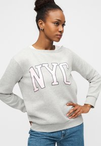 Ανοιχτό γκρι φούτερ με μεγάλα γράμματα "NYC" σε ροζ χρώμα. Απαλή υφή, σχέδιο με λαιμόκοψη και ριμπ μανσέτες. Κανονική εφαρμογή.