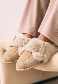 Chaussons beiges avec accents duveteux, tissu texturé, motif chevron et coutures décoratives, ornés de petits points verts et de détails en forme d'oreilles.