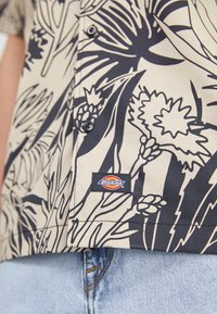 Kremowa koszula z guzikami, z czarnym kwiatowym wzorem, krótkie rękawy i metka Dickies przy dolnym brzegu. Materiał wydaje się gładki i lekki.