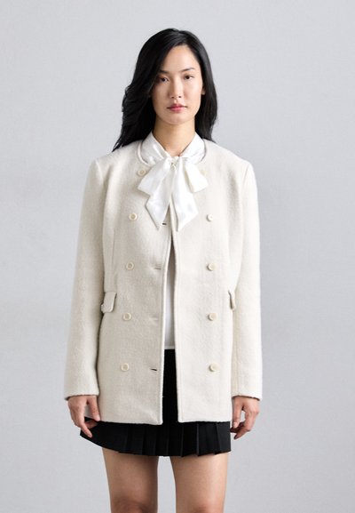Cappotto in lana bianco doppiopetto con tessuto testurizzato, colletto rotondo e bottoni crema, abbinato a una blusa bianca e a una gonna pieghettata nera.