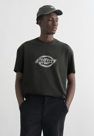 Joven con camiseta negra de Dickies y pantalones oscuros, manos en los bolsillos, mirando hacia un lado, con gorra oscura.