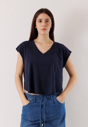 Giovane donna con lunghi capelli castani che indossa una maglia a maniche corte a scollo a V blu navy e pantaloni blu con coulisse, in piedi con le mani nelle tasche.