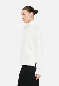 Maglione bianco a collo alto in morbido tessuto a maglia con dettagli a coste, caratterizzato da un piccolo logo a cuore beige all'orlo e spacchi laterali.