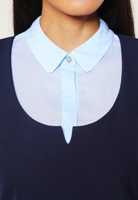 Marineblaues Kleid mit einem hellblauen Button-Down-Kragen, das einen runden Ausschnitt und ein kleines Ausschnittdetail in der Mitte der Vorderseite des Oberteils aufweist.