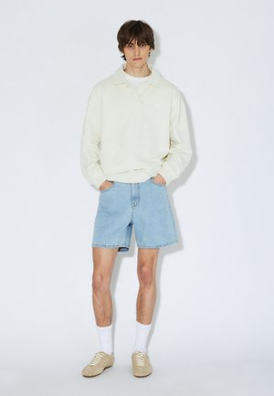 Jasnokremowa bluza z kołnierzem, zestawiona z jasnoniebieskimi szortami dżinsowymi. Beżowe buty i białe skarpetki dopełniają stylizację. Modelka pozuje z rękami w kieszeniach.