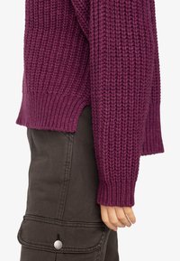 Dicker Strickpullover in dunklem Lila mit gerippten Bündchen, ausgestattet mit einem strukturierten Muster und einem seitlich geschlitzten Saum, kombiniert mit dunklen Cargo-Hosen.