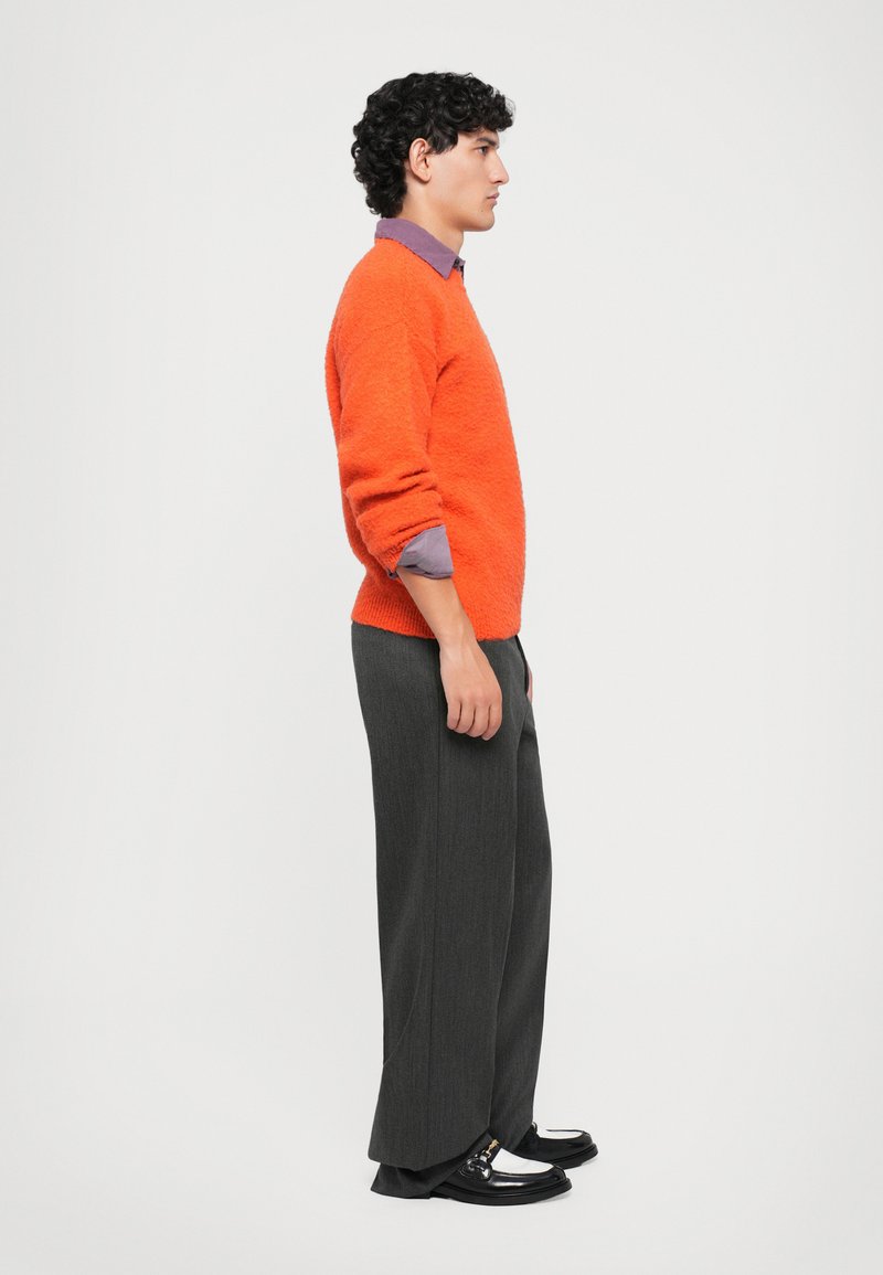 Homme en pull orange, chemise violette, large pantalon gris et mocassins noir et blanc se tient face à droite sur fond blanc.