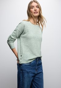 Kobieta z długimi blond włosami ubrana w jasnoszary sweter o teksturze z metalowymi guzikami i luźne niebieskie dżinsy, stojąca na białym tle.