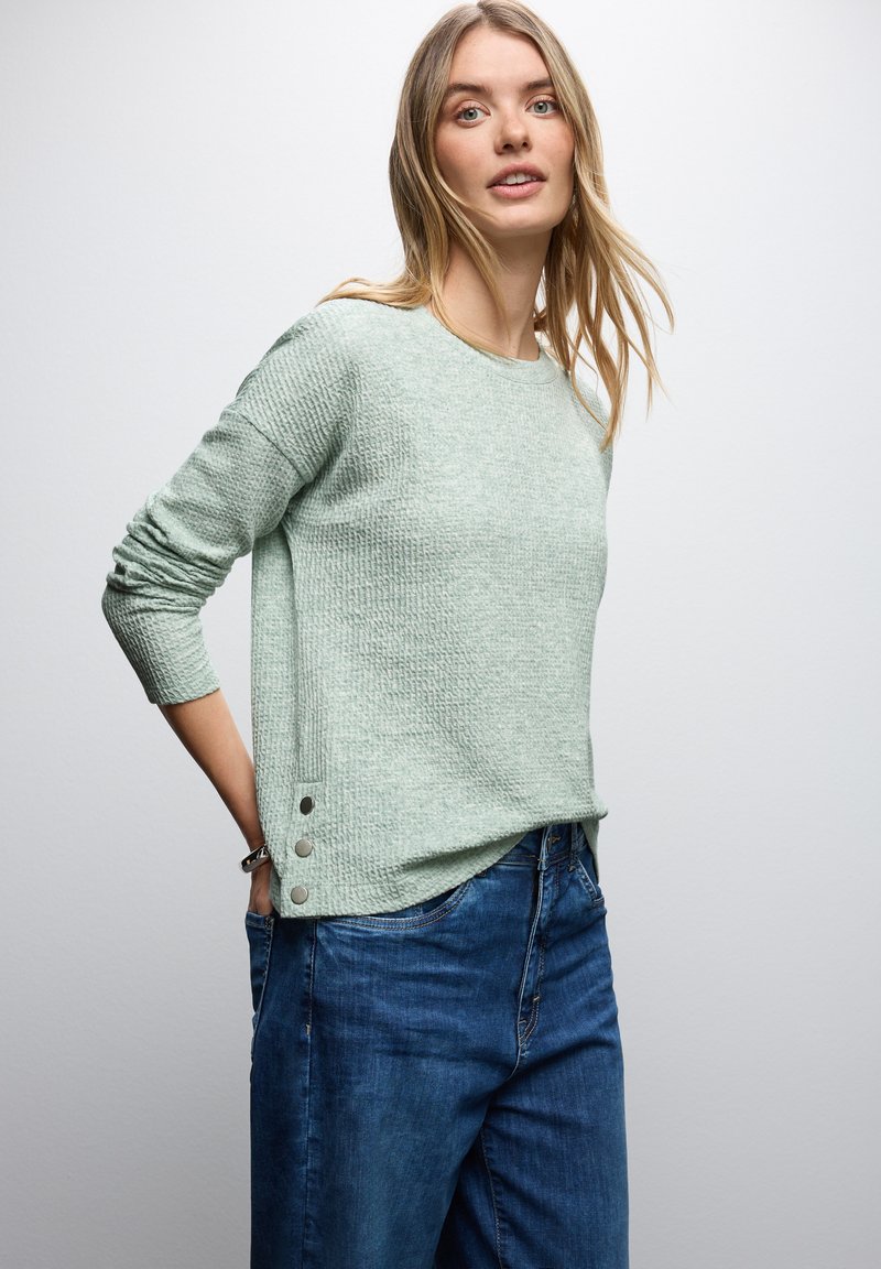 Kobieta z długimi blond włosami ubrana w jasnoszary sweter o teksturze z metalowymi guzikami i luźne niebieskie dżinsy, stojąca na białym tle.