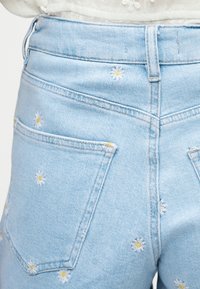 Φωτεινά μπλε τζιν σορτς με κεντημένα λευκά και κίτρινα άνθη μαργαρίτας και εμφανείς πίσω τσέπες.