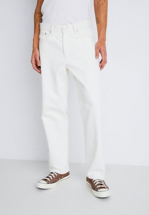 Mężczyzna ubrany w białe jeansy o prostych nogawkach i brązowe niskie trampki, stojący z rękami swobodnie opuszczonymi wzdłuż ciała na jednolitym tle.