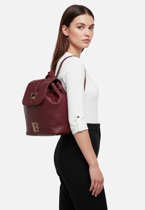 Zaino in pelle color amaranto con parte superiore arrotondata, dettagli in metallo dorato e una lettera "B" prominente sulla parte anteriore. Superficie testurizzata e spallacci regolabili.