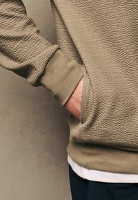 Olivgrüner, strukturierter Pullover mit geripptem Bündchen und Tasche, über einem weißen Hemd getragen. Er hat ein lässiges, lockeres Design.