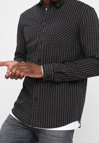 Koszula na guziki w czarno-szare kratę, z długimi rękawami, pojedynczą kieszenią na piersi i zaokrąglonym dołem, wykonana z lekkiego materiału.