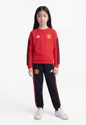 Młoda dziewczyna stoi, ubrana w czerwoną bluzę Manchester United i czarne spodnie dresowe z czerwonymi paskami oraz białe trampki, zwrócona przodem.