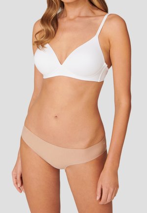 Frau trägt einen weißen drahtlosen BH und beige nahtlose, niedrig geschnittene Slips vor einem schlichten hellgrauen Hintergrund.