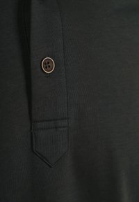 Donkergrijze stof met een gladde textuur heeft een horizontale pocket en een bruine knop met vier gaten.