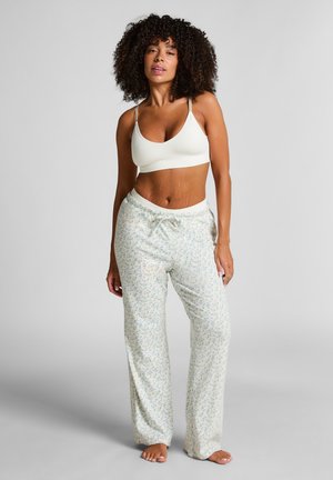 Woman with curly hair wearing a white crop top and light-colored floral pajama pants standing barefoot on a gray background.