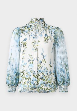 Blouse blanche avec imprimé floral bleu, col montant à volants, manches longues plissées transparentes et poignets smockés.