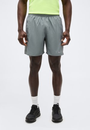 Άνδρας ντυμένος με γκρι αθλητικά σορτς, φανέλα νέον πράσινου χρώματος, λευκές κάλτσες και μαύρα αθλητικά παπούτσια, στέκεται μπροστά σε ένα απλό φόντο.