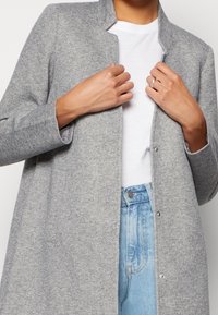 Cardigan largo gris texturizado con cuello alto, con cierres de broche. Combinado con una camiseta blanca y vaqueros azul claro.
