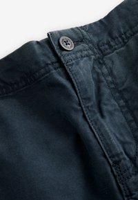 Mørkeblåt bomuldsstof med en glat tekstur. Har en sort knap ved taljen med præget branding og fine syningsdetaljer.