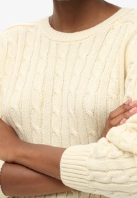 Cream cable knit sweater featuring a textured pattern, ribbed cuffs, and a classic round neckline. Material appears soft and cozy.