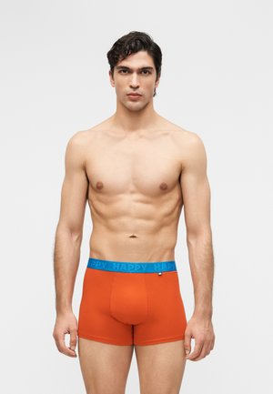 Mladý muž s tmavými vlasmi v oranžových boxerkách s modrým pásom označeným slovom "HAPPY", stojaci na jednoduchom sivom pozadí.