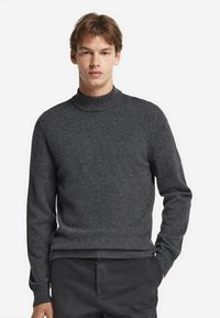 Junger Mann in einem dunkelgrauen Rollkragensweater und schwarzen Hosen, der mit einer Hand in der Tasche vor einem schlichten weißen Hintergrund steht.