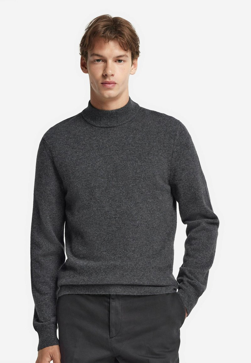 Junger Mann in einem dunkelgrauen Rollkragensweater und schwarzen Hosen, der mit einer Hand in der Tasche vor einem schlichten weißen Hintergrund steht.