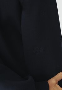 Sweatshirt bleu marine avec une texture douce, orné de motifs de ours en relief subtils sur les manches, illustrant un design classique et décontracté.