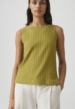 Frau trägt ein ärmelloses, geripptes olivgrünes Oberteil und weite weiße Hosen, steht mit einer Hand in der Tasche vor schlichtem Hintergrund.