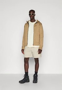 Felpa con zip color ruggine, t-shirt crema, pantaloncini beige, stivali da trekking neri, accessoriata con occhiali da sole bianchi. Texture lisce, design casual.