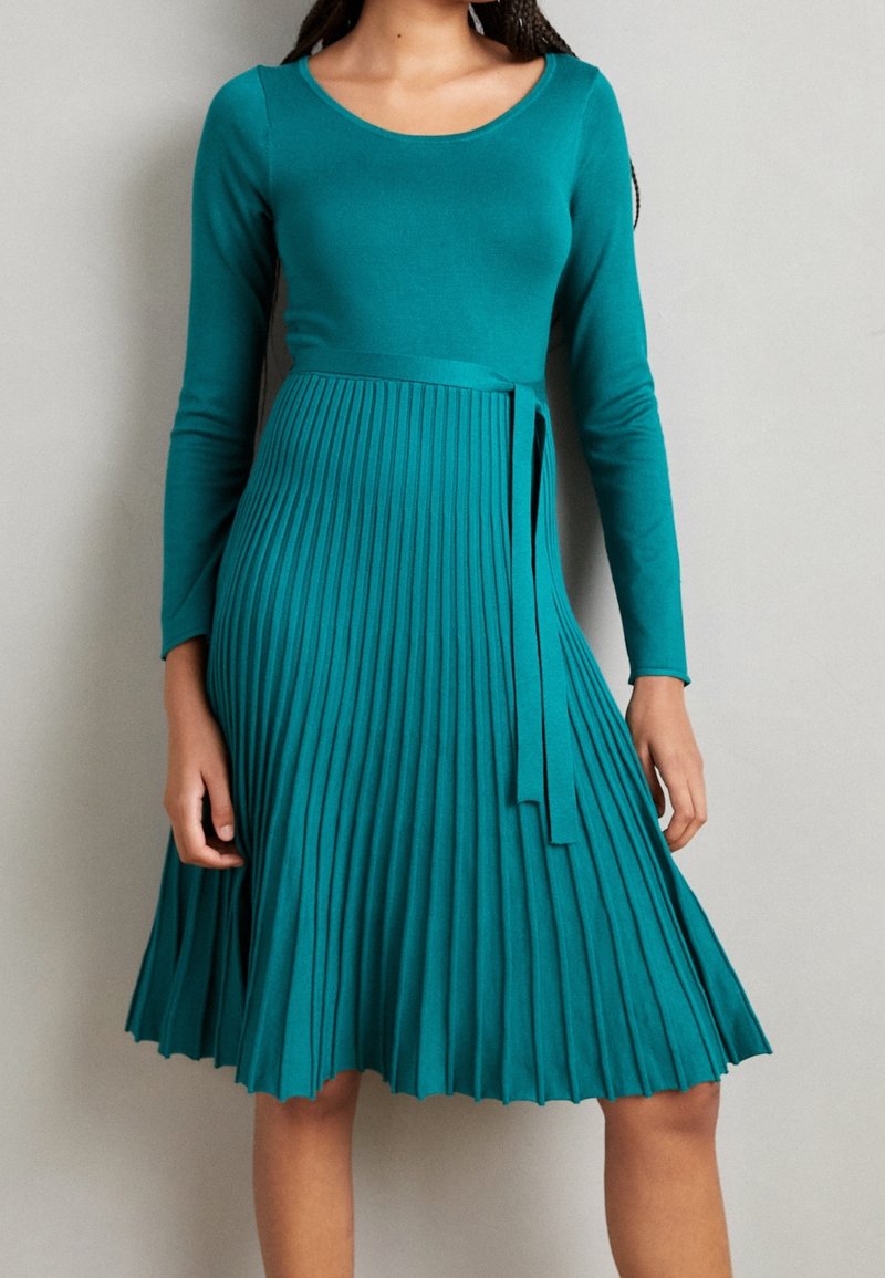 Femme portant une robe bleu canard à manches longues, haut ajusté, jupe plissée et ceinture assortie, debout devant un fond clair et uni.