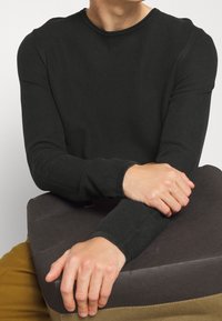 Homme portant une chemise noire texturée à manches longues, reposant les bras croisés sur une surface rembourrée sombre, avec un pantalon moutarde visible.