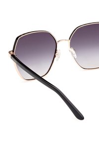 Gafas de sol negras y doradas con un marco hexagonal y lentes grises degradados. Cuentan con patillas texturizadas en negro y almohadillas nasales transparentes.