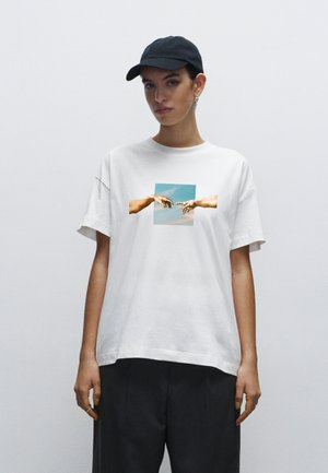Witte katoenen t-shirt met een graphic van twee handen die naar elkaar reiken tegen een blauwe lucht achtergrond. Ontspannen pasvorm.