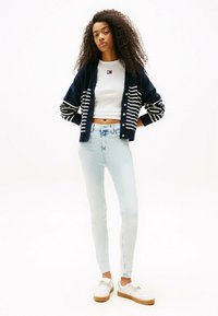 Cárdigan a rayas azul marino con acentos blancos sobre una blusa blanca corta, combinado con vaqueros ajustados de color azul claro y zapatillas blancas con suela beige.